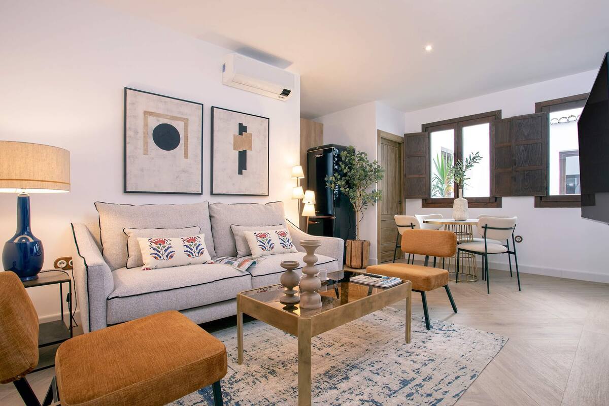 Ein modernes Wohnzimmer mit Sofa, Couchtisch und Sesseln. Es gibt eine Essecke mit Tisch und Stühlen. An den Wänden befinden sich Kunstwerke und eine Pflanze.