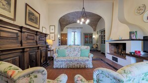 A spacious living room with a floral sofa, a wooden cabinet, and a fireplace. - Il Cerreto 11 Emma Villas (Pontedera)