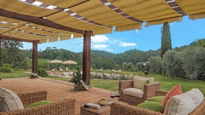 A covered outdoor seating area with wicker furniture and umbrellas, overlooking a lush garden and distant hills. - Il Cerreto 11 Emma Villas (Pontedera)