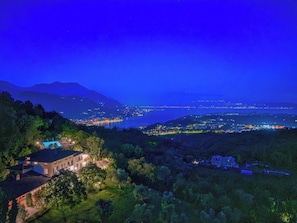 Vista notturna di una zona costiera con un ampio specchio d'acqua, montagne sullo sfondo e un edificio con piscina in primo piano.