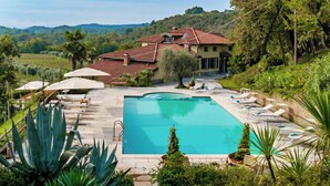 Un espace piscine agrémenté de chaises longues et de parasols, entouré d’une végétation luxuriante et d’une maison dont le toit est recouvert de tuiles rouges.