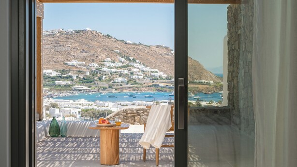 A balcony with a table, a chair, and a view of a coastal town with buildings and boats.