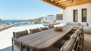 A wooden dining table with chairs, a bowl of fruit, and a view of a swimming pool and buildings in the distance.
