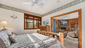 A bedroom with a large bed, a wooden headboard, a window with blinds, a wall-mounted TV, and a view of the outdoors. - Cozy Mountain Retreat • Near RMNP & Downtown (Estes Park)