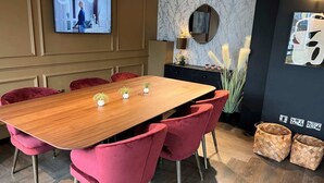 A conference room with a wooden table, red chairs, a TV on the wall, a black cabinet with decorative items, and a black wall with a framed picture. - The Depot in Bath (Bath)