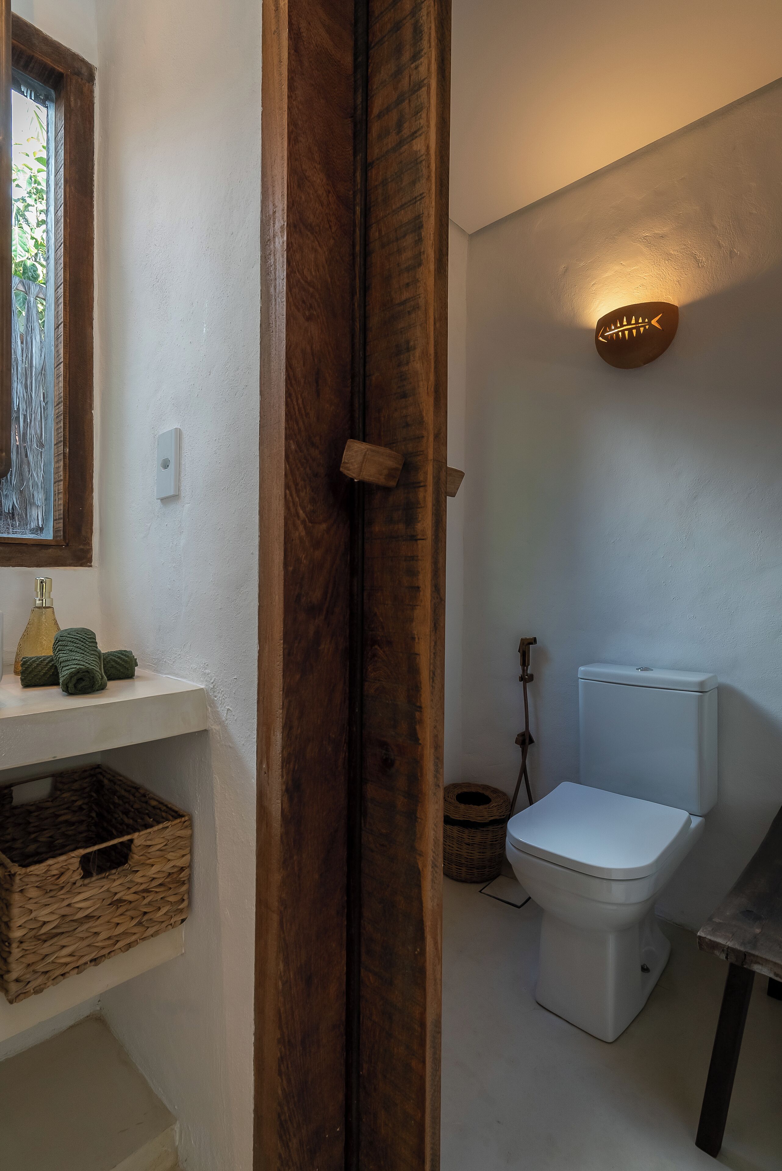 A bathroom with a white toilet, a wooden door, a shelf with baskets, and a wall-mounted light fixture.
