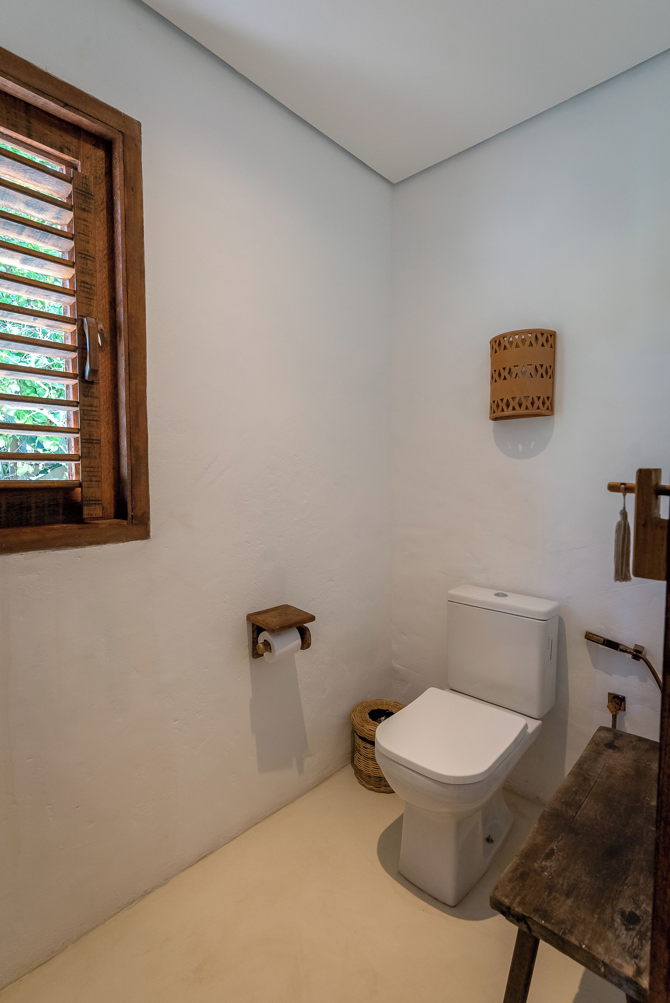 A small bathroom with a toilet, a wooden shelf, a wicker basket, and a window with blinds.