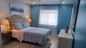 A bedroom with a tufted headboard bed, patterned bedding, a nightstand with a lamp, a window with sheer curtains, and a vase on a side table. - Modern 2BD/BA, 1.5 Blocks to Sea, Walk to Amenities w/ Pool, Linens & Parking (Virginia Beach)