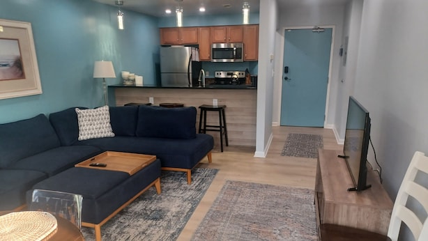 A modern living room with a dark blue sofa, a wooden coffee table, and a TV on a stand. - Modern 2BD/BA, 1.5 Blocks to Sea, Walk to Amenities w/ Pool, Linens & Parking (Virginia Beach)