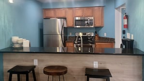A kitchen with wooden cabinets, stainless steel appliances, and a black countertop. There are two stools and a fire extinguisher mounted on the wall. - Modern 2BD/BA, 1.5 Blocks to Sea, Walk to Amenities w/ Pool, Linens & Parking (Virginia Beach)