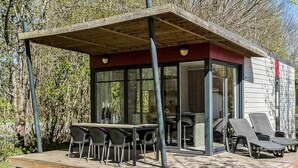 A modern outdoor dining area with a table and chairs, a covered seating area with lounge chairs, and a glass-enclosed structure with a red accent wall. - 6 Person Holiday Park Home in Hesselager (Hesselager)