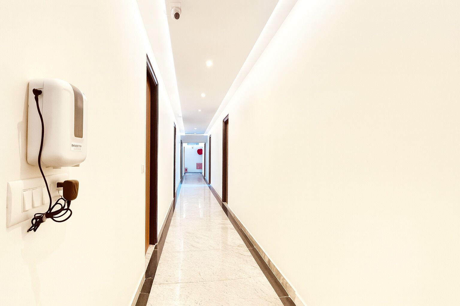 A hotel corridor with a hand sanitizer dispenser, a power outlet, and a red circular sign on the wall.