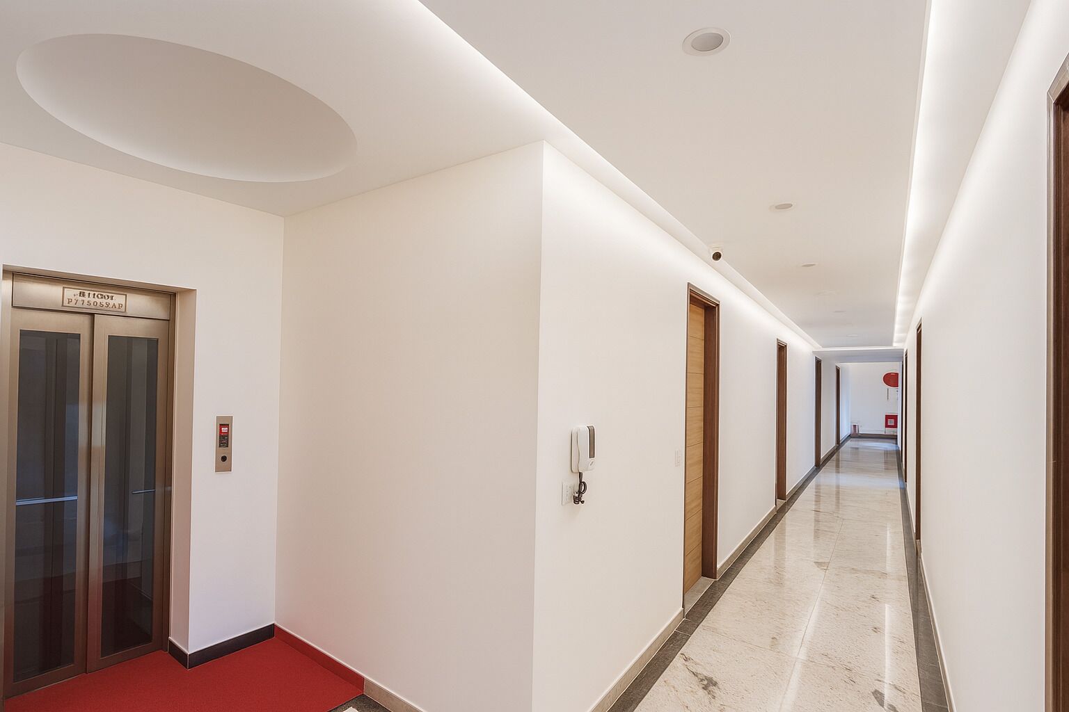 A modern hotel corridor with a red carpet, an elevator, and a long hallway with doors and a fire alarm.