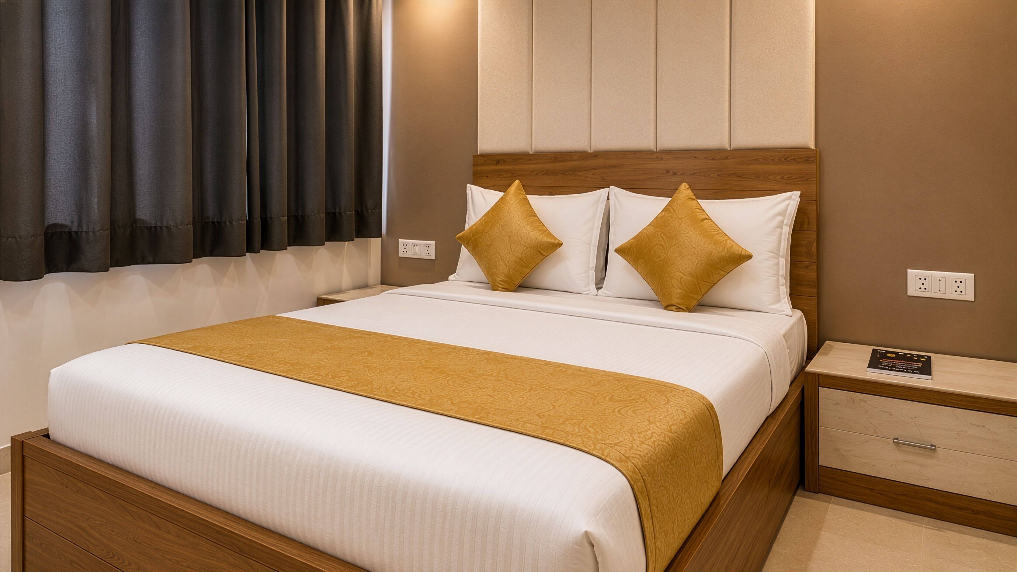 A hotel room with a bed, a nightstand, a drawer, a window with curtains, and a wall socket.