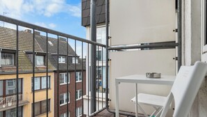 Een balkon met een witte tafel en stoel, een metalen hekwerk en uitzicht op omliggende gebouwen.