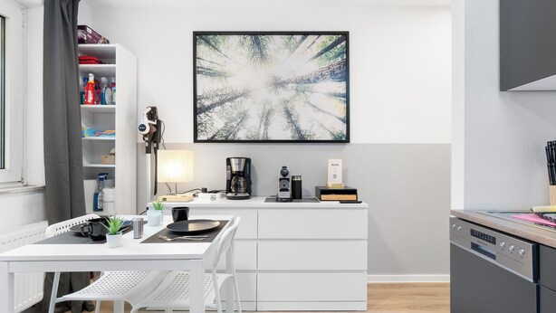 Een moderne keuken met witte kastjes, een eettafel met stoelen, een koffiezetapparaat en een groot abstract schilderij aan de muur.