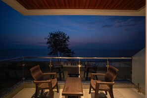 Varanda com vista para o oceano ao entardecer, com móveis de madeira e guarda-corpo de vidro.