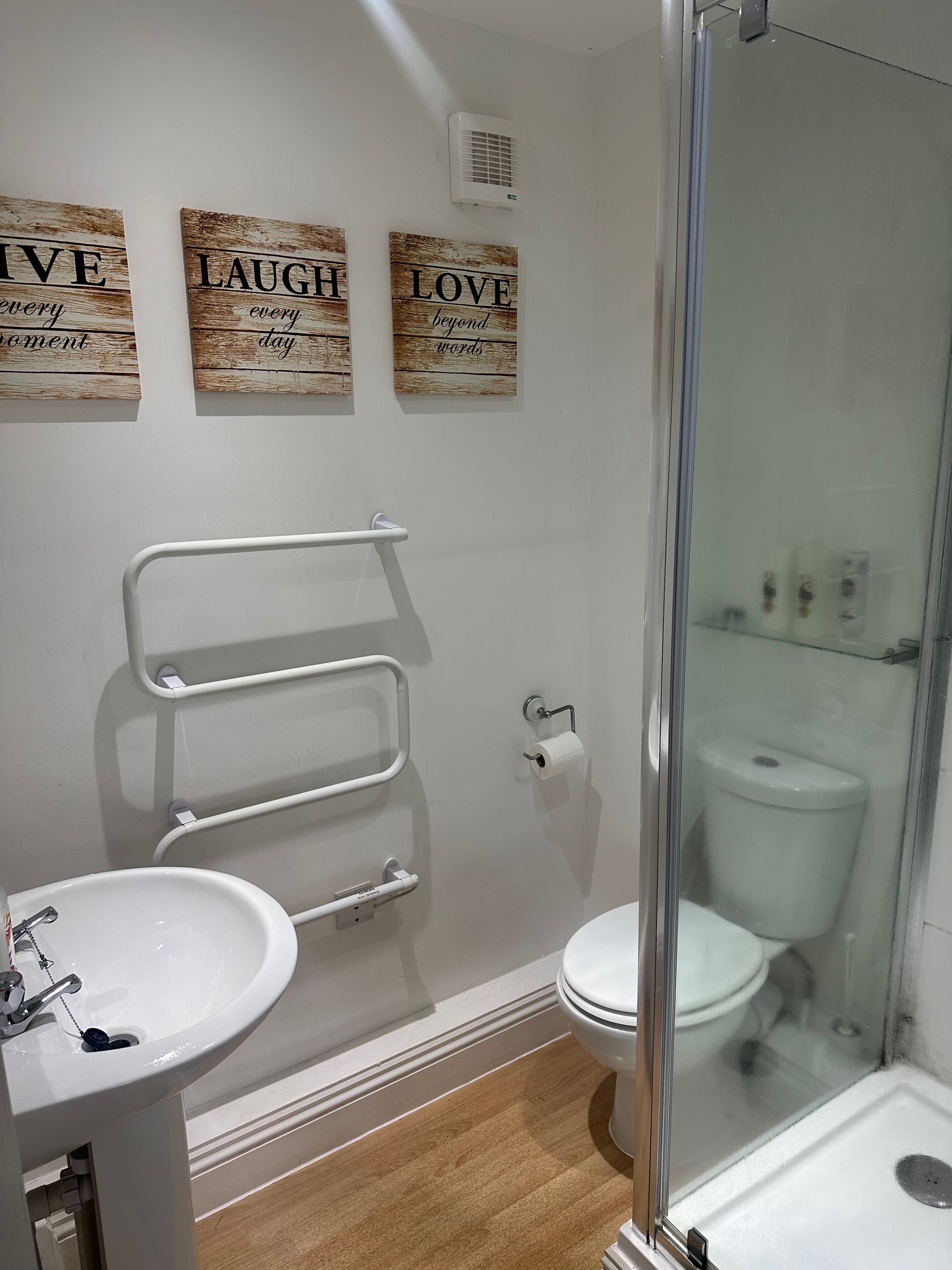 A bathroom with a sink, toilet, and glass shower enclosure. There are three wooden signs with motivational phrases on the wall.