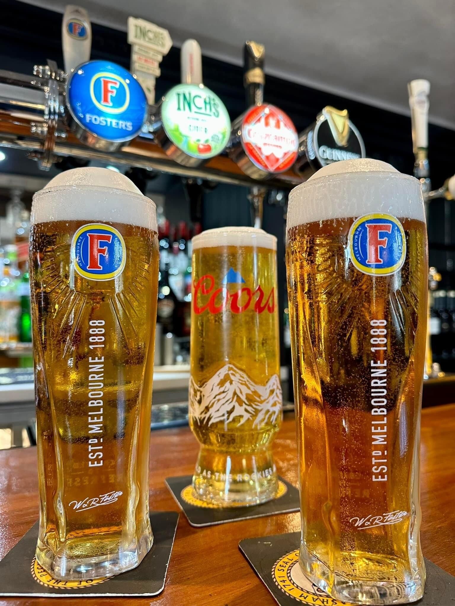 Tres vasos altos de cerveza con logotipos y texto, sobre posavasos en un bar, con una grifería de fondo.