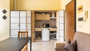 Een kleine keuken met een koelkast, magnetron en wastafel. Er is een eettafel met stoelen en een bank met kussens.