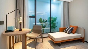 Ein modernes Zimmer mit einer Couch, einem kleinen Tisch und einem Sessel. Ein großes Fenster mit Vorhängen bietet Blick auf die grüne Umgebung.