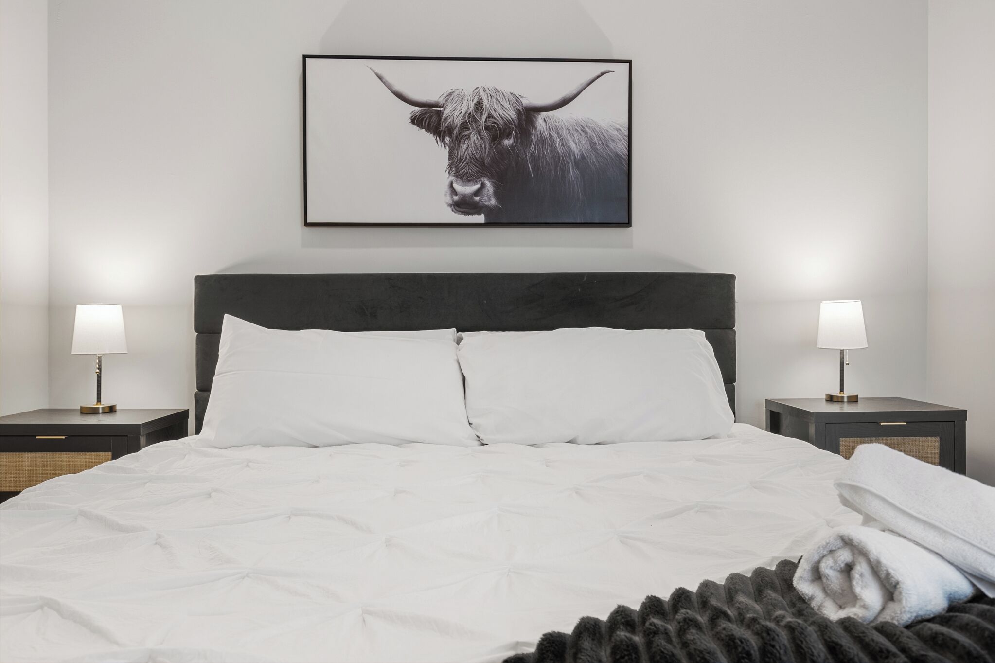 Een keurig opgemaakt bed met witte lakens, twee nachtkastjes met lampen, en een ingelijst schilderij van een stier aan de muur.