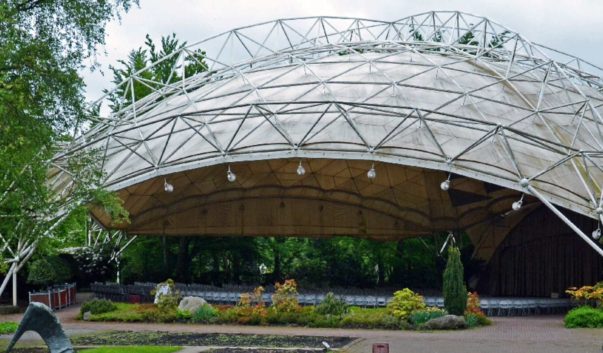 A large, semi-circular, metal-roofed structure with a grid-like pattern, surrounded by landscaped gardens and trees.