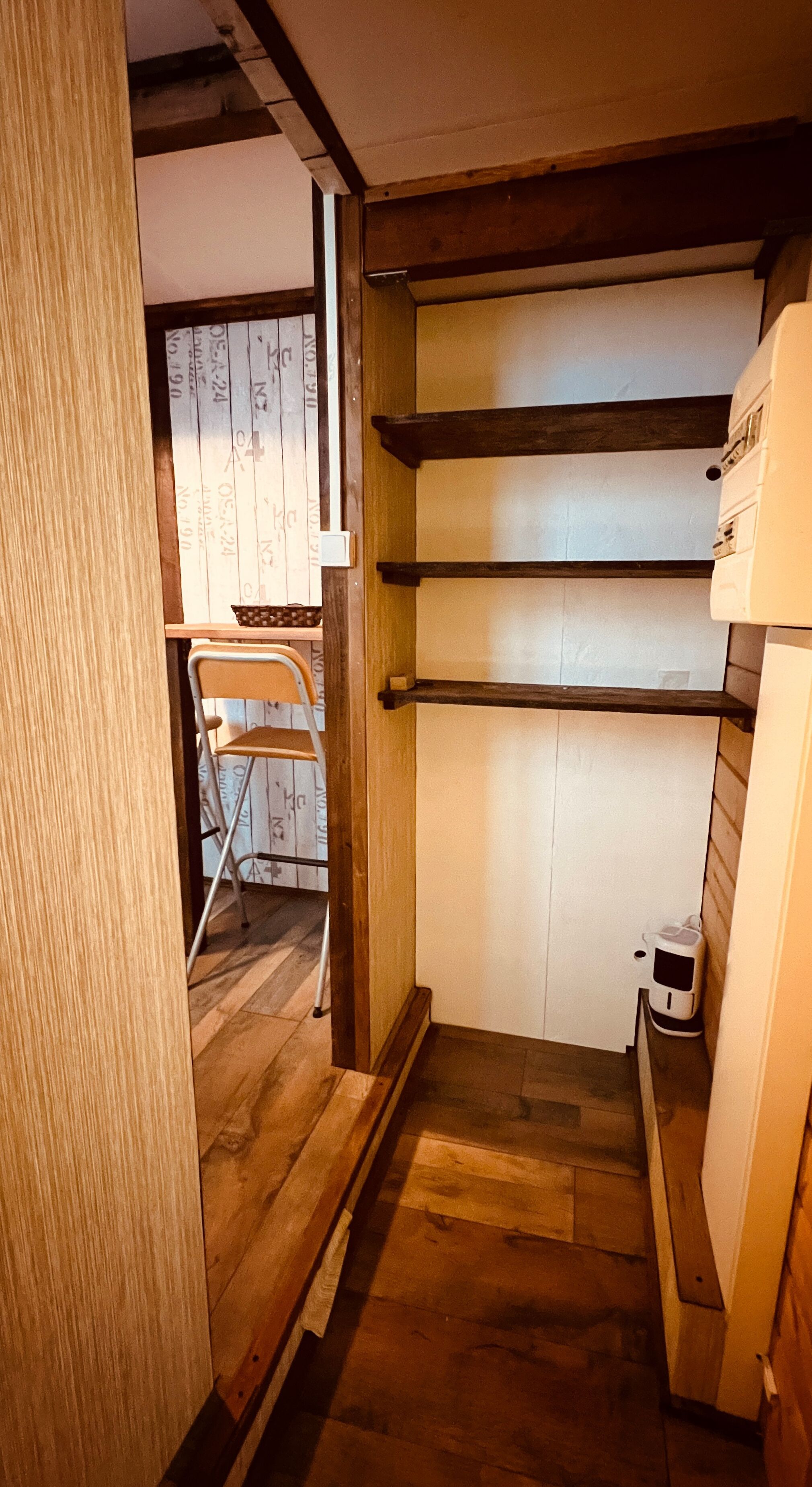 A narrow hallway with wooden flooring and a white door. Shelves are mounted on the wall, and a small white appliance is visible on the right.