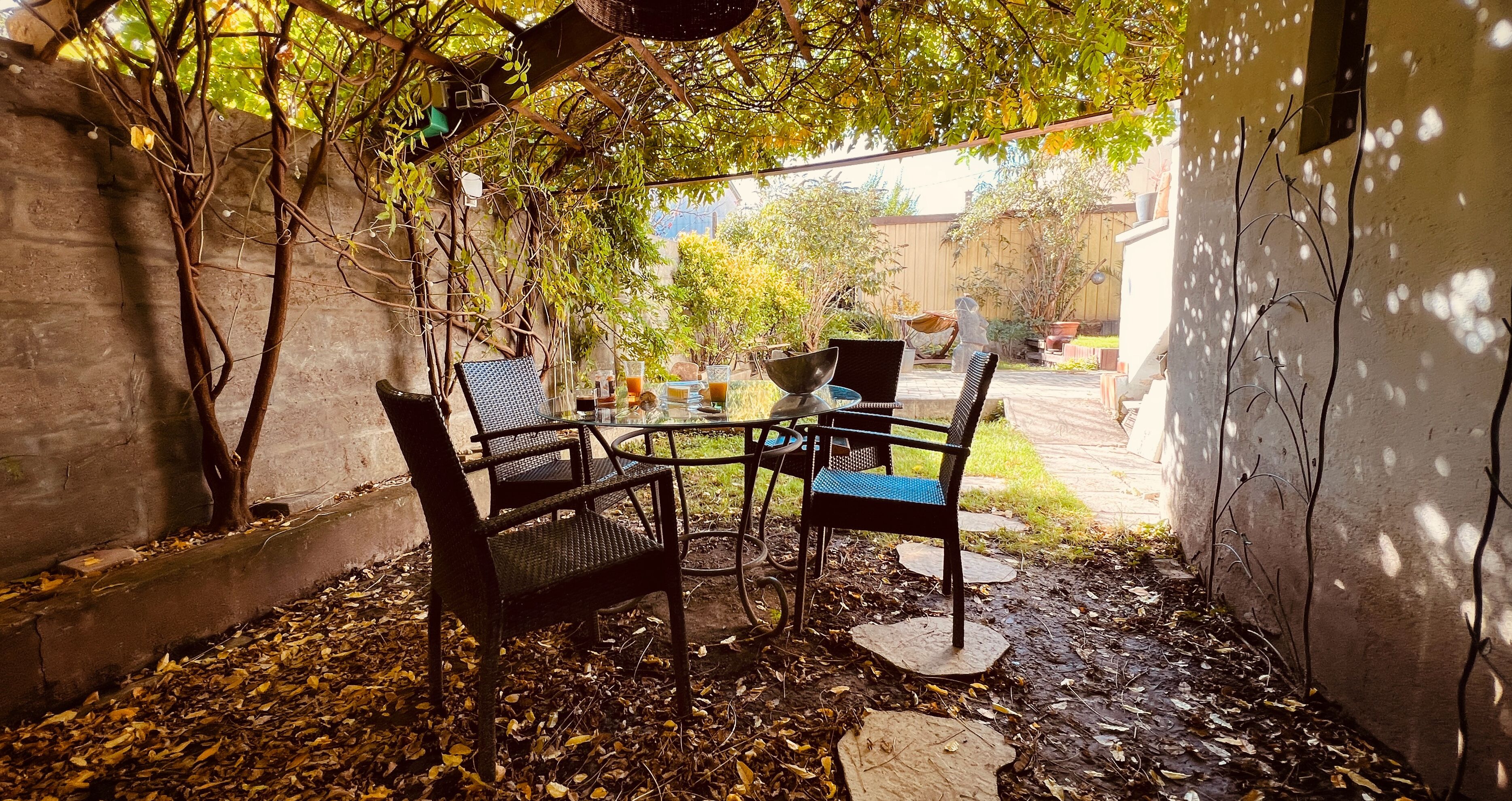 An outdoor seating area with metal chairs and a table, surrounded by trees and a concrete wall.