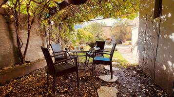 An outdoor seating area with metal chairs and a table, surrounded by trees and a concrete wall.