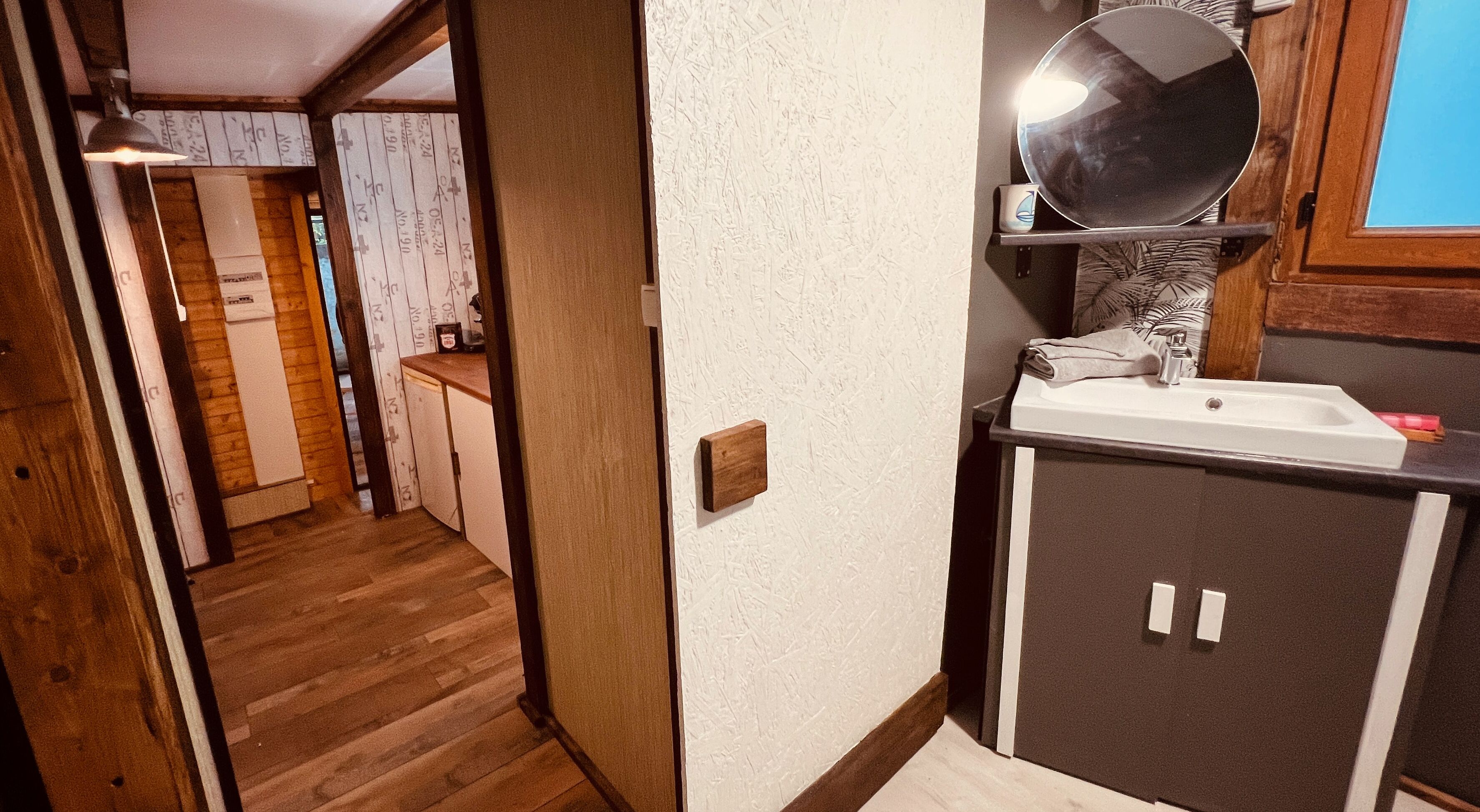 A narrow hallway leading to a kitchen area with a wooden floor, a white sink, and a mirror above it.