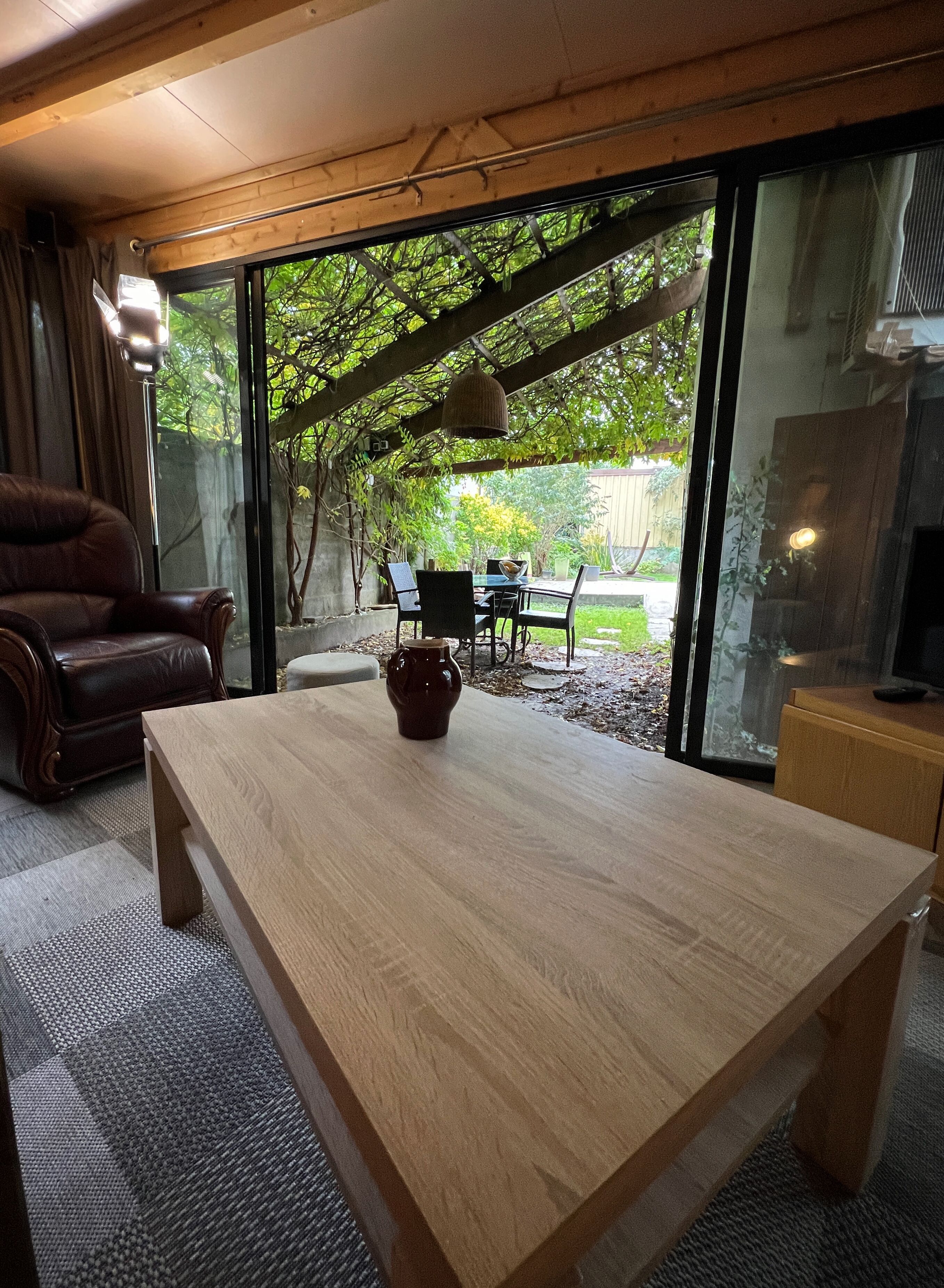 Une table à manger en bois, ornée d’un vase au centre, un canapé en cuir et une porte en verre donnant sur un espace extérieur agrémenté d’une terrasse et de verdure.