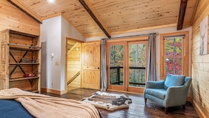 Interno di una baita in legno con un letto, una sedia, un tappeto e una scala che conduce a un balcone.