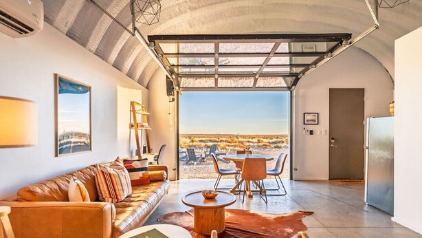 Ein geräumiges Wohnzimmer mit einer Ledersofa, einem Couchtisch und einem Esstisch mit Stühlen. Ein großes Fenster bietet Blick auf die Wüstenlandschaft.