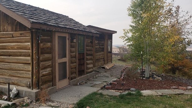 Une cabane rustique en rondins de bois, avec une façade en bois, un porche et un petit jardin agrémenté d’un arbre et de rochers.