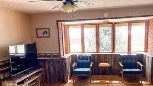 Un salon chaleureux avec un plancher en bois, un téléviseur posé sur un meuble en bois, deux fauteuils en cuir bleu, une petite table ronde et une grande fenêtre donnant sur la forêt.