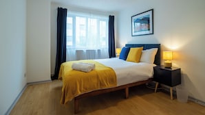 A neatly made bed with a yellow bedspread and pillows, a bedside table with a lamp, and a window with curtains. - Captivating 3-bed House in London (London)
