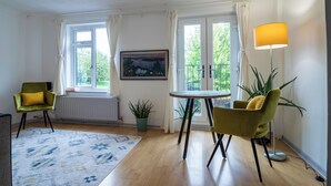 A room with two green armchairs, a small round table, a floor lamp, a radiator, a window with a view of greenery, and a painting on the wall. - Captivating 3-bed House in London (London)