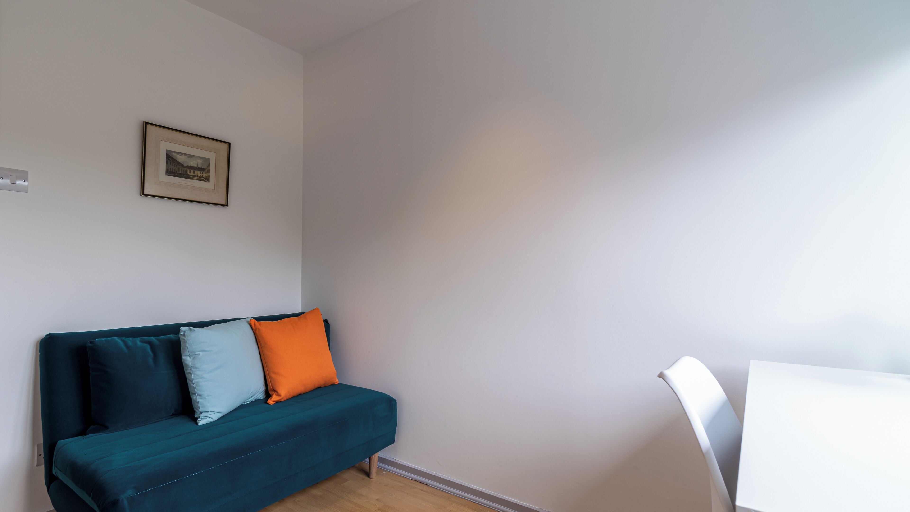 Un salon minimaliste avec un canapé de couleur turquoise, deux coussins, un tableau encadré accroché au mur et une table blanche.