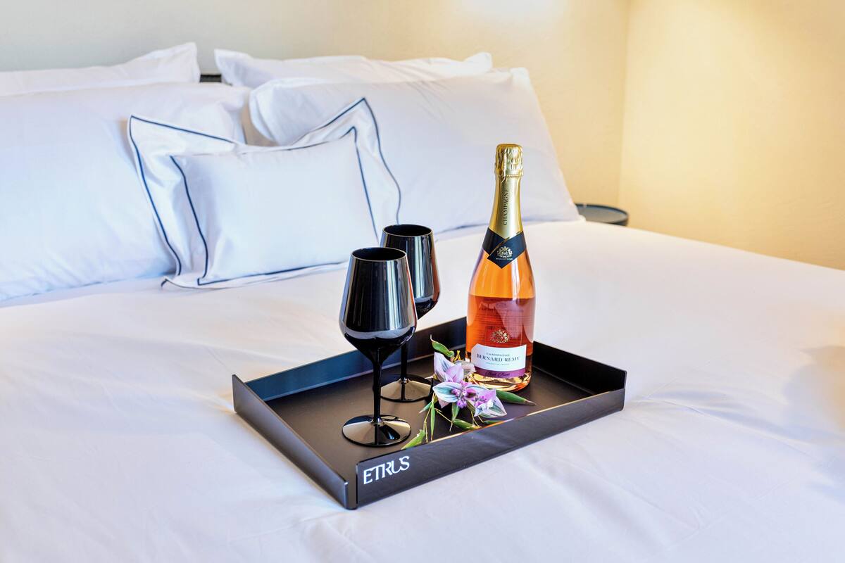 A bed with white linens and pillows, a tray with a bottle of champagne, two glasses, and a flower.
