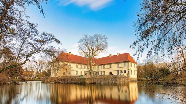 A large, historic building with a red-tiled roof, situated by a calm body of water, surrounded by trees. - RtR Herne (Herne)