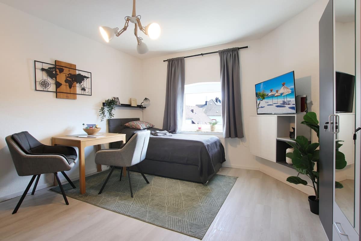 A modern bedroom with a bed, desk, chairs, a television, and a window with curtains.