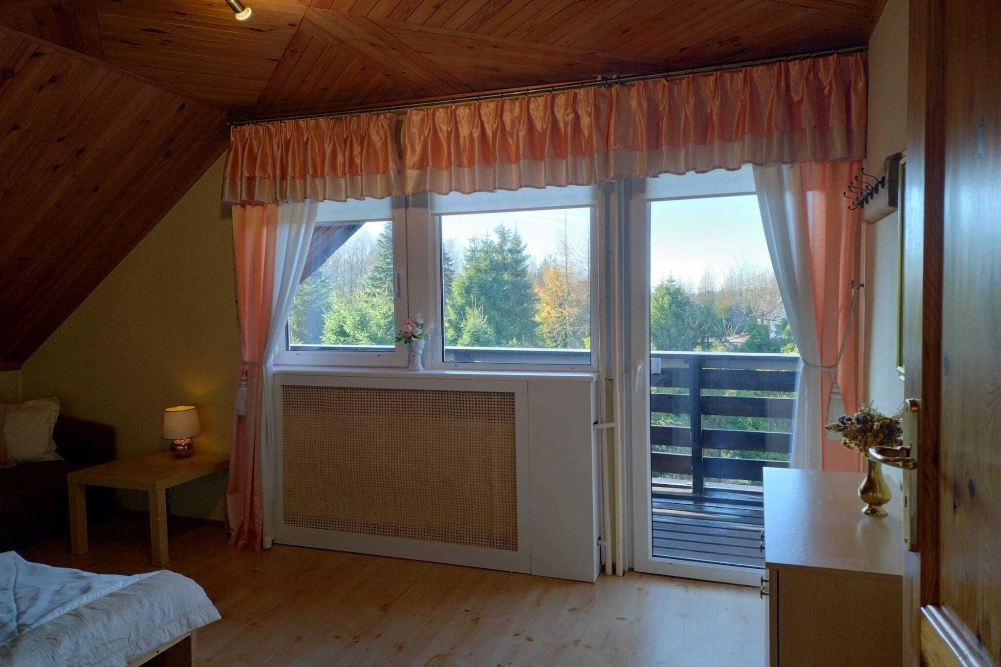 Une pièce avec un plafond en pente, un lit, une petite table avec une lampe, une fenêtre avec des rideaux et une vue sur les arbres à l’extérieur.