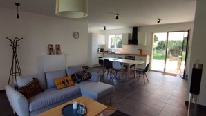 A modern living room with a sofa, coffee table, and lamp. A kitchen with a dining area is visible in the background. - WEST COAST house between ocean, Bassin d'Arcachon, Dune du Pyla, Bordeaux (Le Porge)