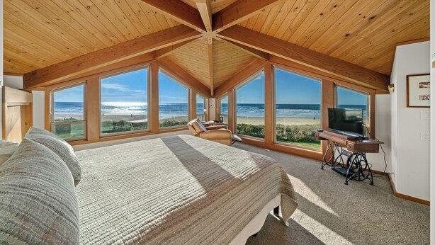 Ein Schlafzimmer mit einem großen Bett, einem Fernseher auf einem Holztisch und Blick auf den Strand durch große Fenster.