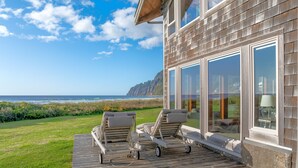 Eine Holzterrasse mit zwei Liegestühlen und einem Tisch mit Blick auf einen Strand und steile Klippen.