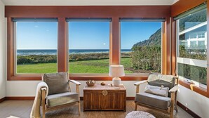 Ein Zimmer mit großen Fenstern, einem Holztisch, zwei Sesseln und einer Lampe.