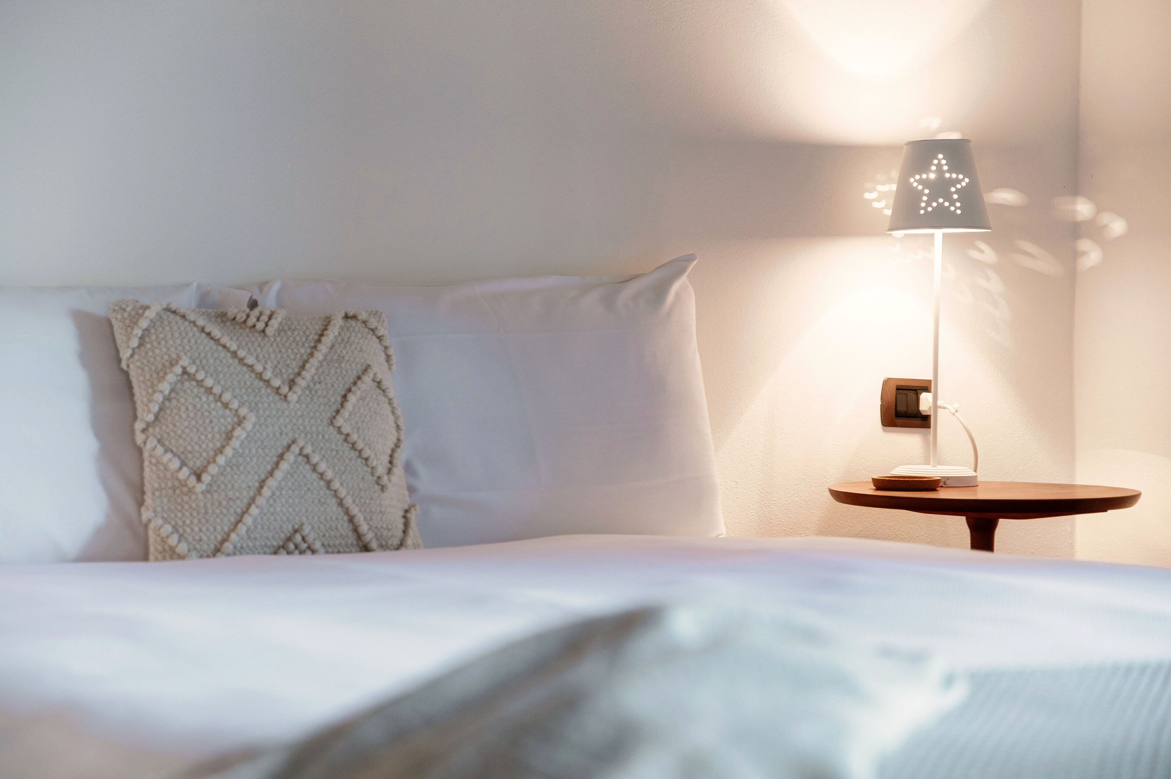 Een net opgemaakt bed met een wit kussen en een deken met een patroon. Er staat een klein, rond tafeltje met een lamp met een stervormige kap.