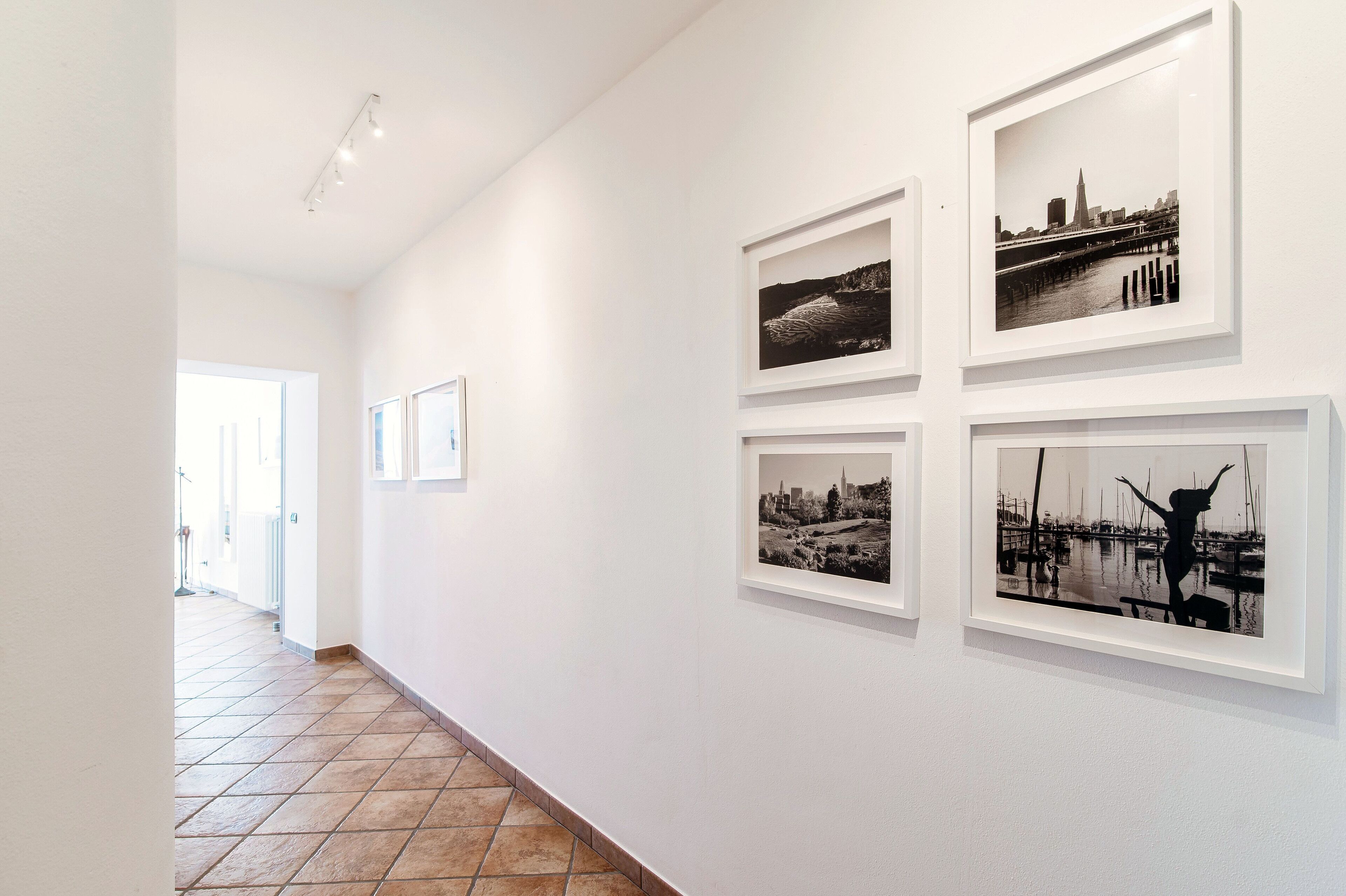 A hallway with framed black and white photographs on the wall, leading to a bright room with a tiled floor.