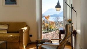 A living room with a sofa, a chair, a table, and a floor lamp. There is a balcony with a view of mountains and trees.